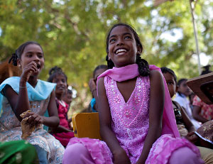 Young smiling Sri Lankan girls in bright coloured dresses sitting down. Behind them are a number of other children sitting down.