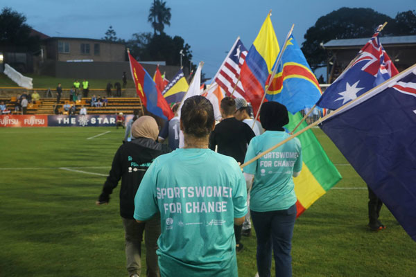 People walking onto a field each holding a country flag. 3 Women wearing sports women for change jerseys in the centre of the photo. 