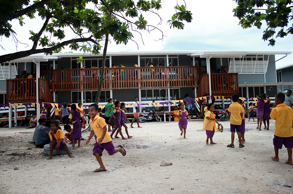 Children in brightly coloured school uniforms run and play on the sand, with a school building in the background.