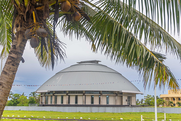 A circular building with a silver domed roof sits on a raised foundation, with a coconut tree with coconuts in front of it.