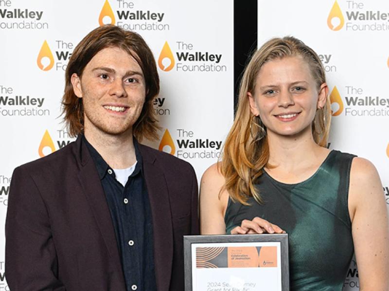 Man and a women standing in front of a Walkley Foundation media wall, holding a framed certificate.