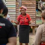 Woman wearing a red shirt and flower headpiece speaks to a group of people. 