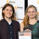 Man and a women standing in front of a Walkley Foundation media wall, holding a framed certificate.