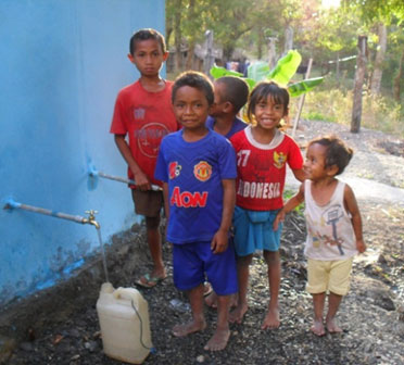 Five young children and standing beside a blue wall with a bucket besides them being filled up from a tap coming out of the wall.