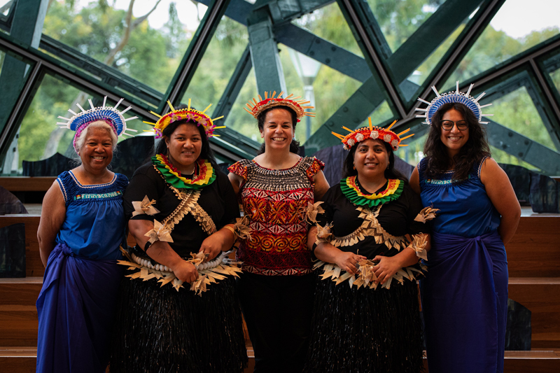 Five women standing together smiling, wearing traditional indigenous clothing and headwear.