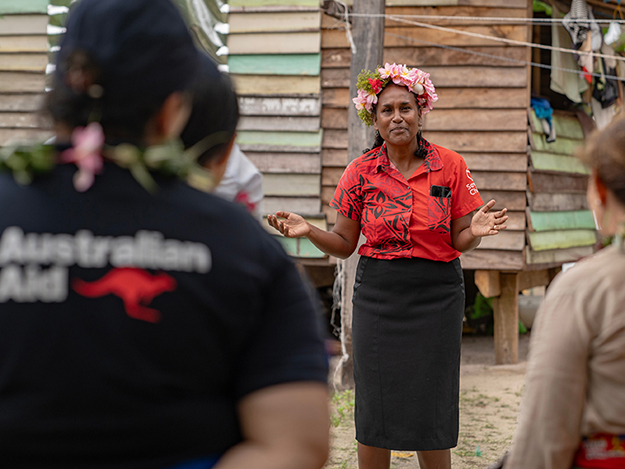 Woman wearing a red shirt and flower headpiece speaks to a group of people. 