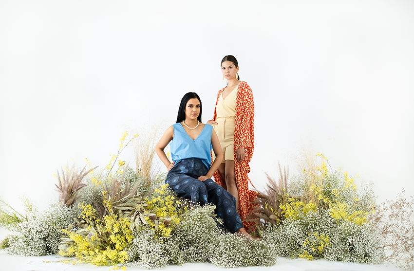 front model sitting down in a blue, sleeveless top and blue trousers, second model standing to the right in beige shorts and t-shirt with a long, orange, flowy cardigan. Copious amounts of arranged fauna on the ground.