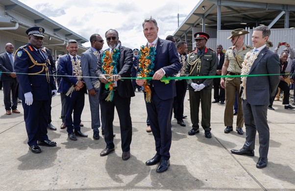 Two men standing together cutting a green ribbon. Men in military uniforms of Vanuatu and Australia, and other dignitaries stand by.