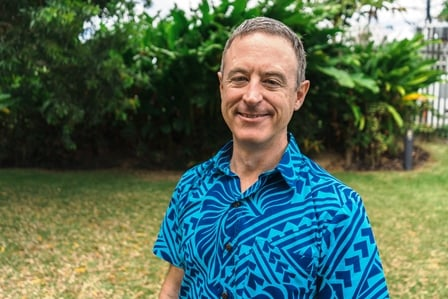 A man in a bright, blue, patterned shirt standing outside, smiling