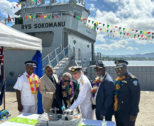 Two men cutting into a cake, shaped like a naval ship, as others in naval uniform stand by. The RVS Sokomanu is in the background, decorated in colourful bunting.